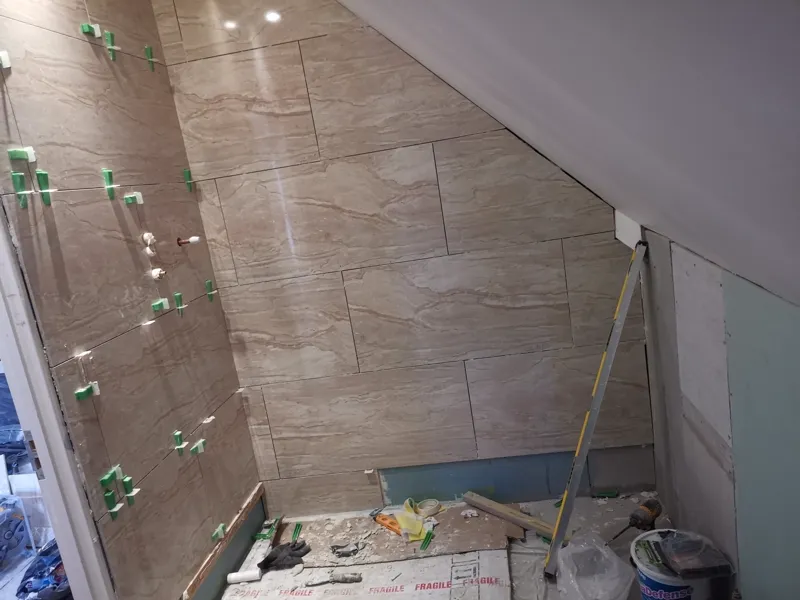 Large-format tiles being fixed to a sloped loft-eaves wall and ceiling, spacers in place, grout bucket on the floor.