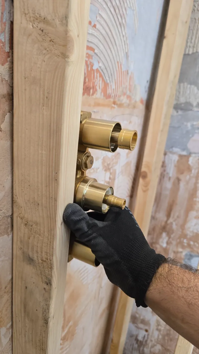 A gloved hand fitting a brass shower valve body between timber studs.
