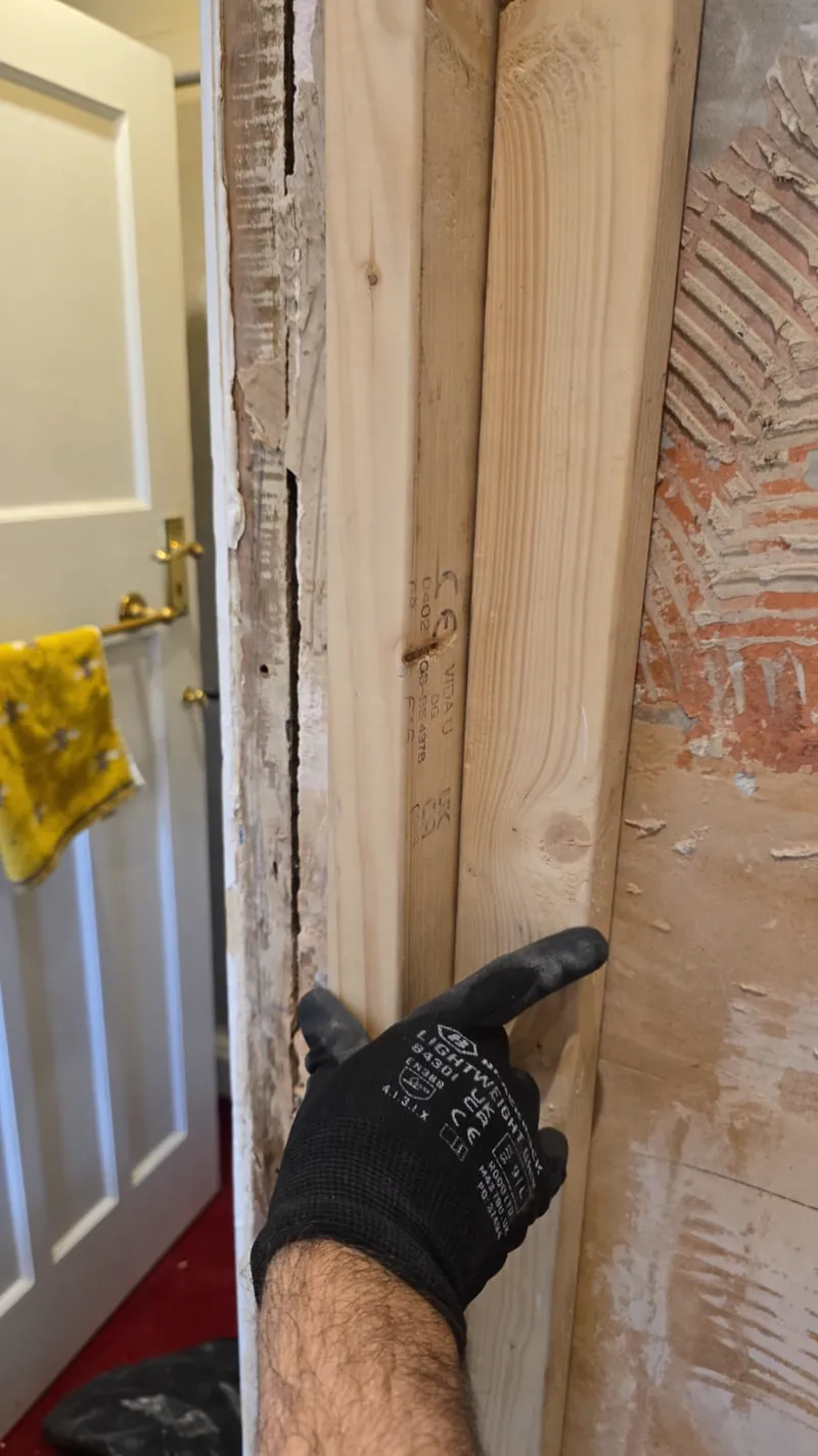 A gloved hand pointing to a timber door-frame repair beside a plastered wall.