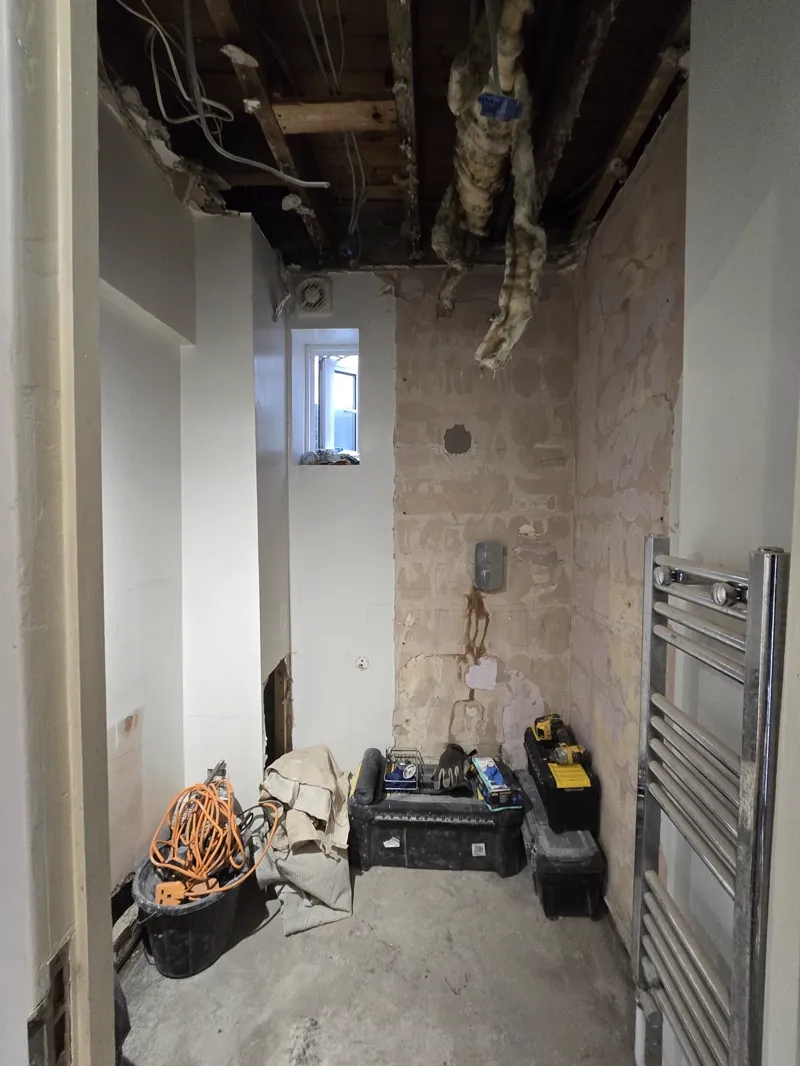 Bathroom mid strip-out with a heated towel rail, tool boxes and cabling on the floor, ceiling timbers exposed above.