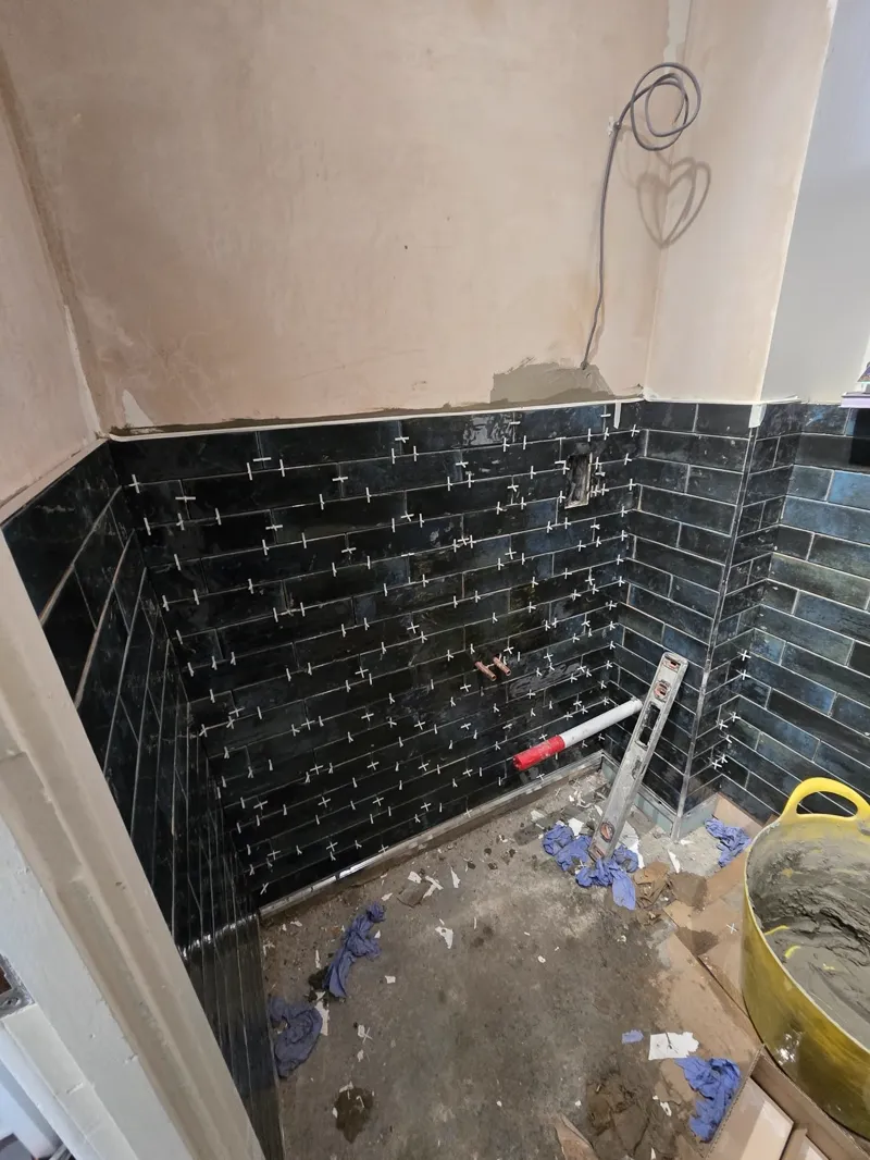 Black metro tiles installed on bathroom walls, spacers still in place, tools and materials on the floor.