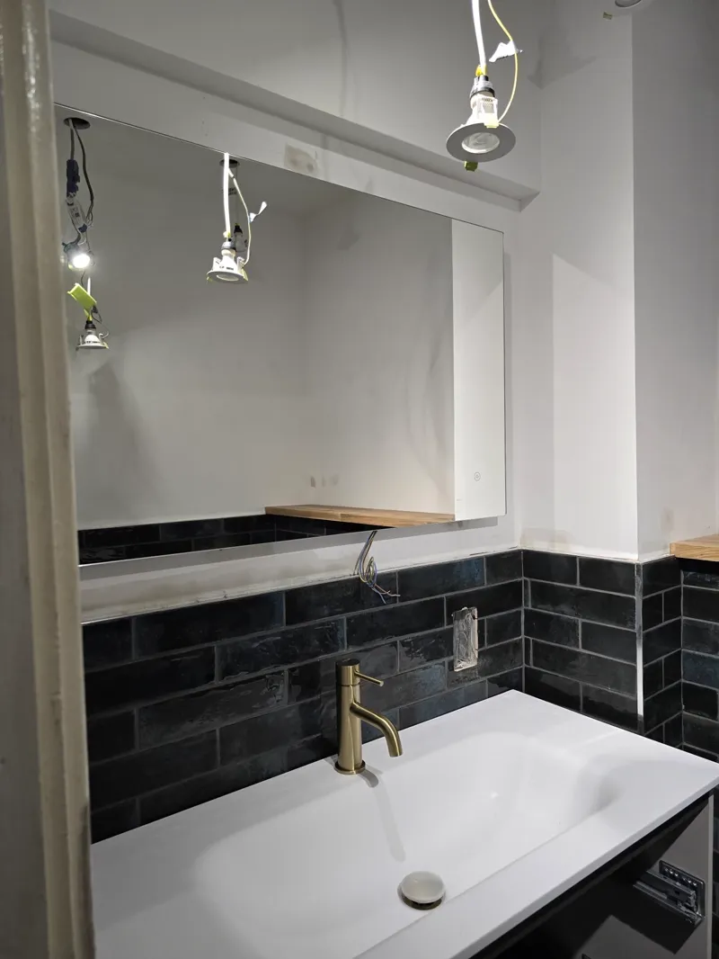 A large mirror above a black metro-tiled splashback with a white countertop basin and brass mixer tap.