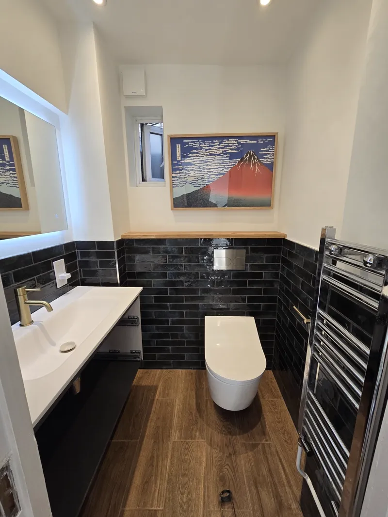 Completed cloakroom with black metro-tile walls, a wall-hung toilet, a framed print and a chrome heated towel rail.